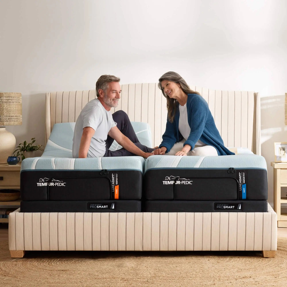 Two people sitting on a Tempur-Pedic mattress in a bedroom setting.